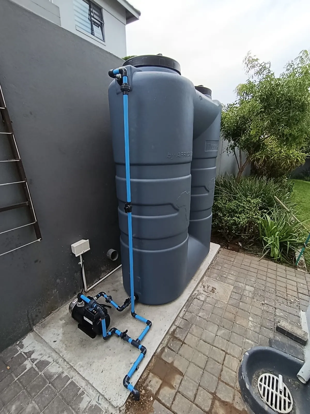 Two slimline water tanks with a pump and blue pipework against a house wall